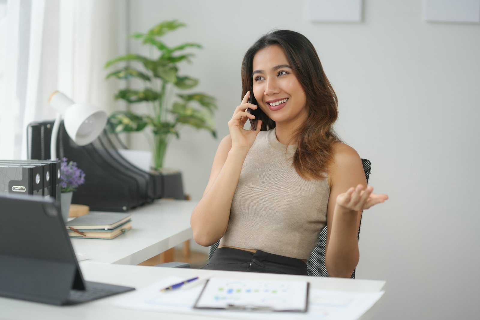 Woman smiling while talking on the phone at her desk in a modern office with documents and a laptop.